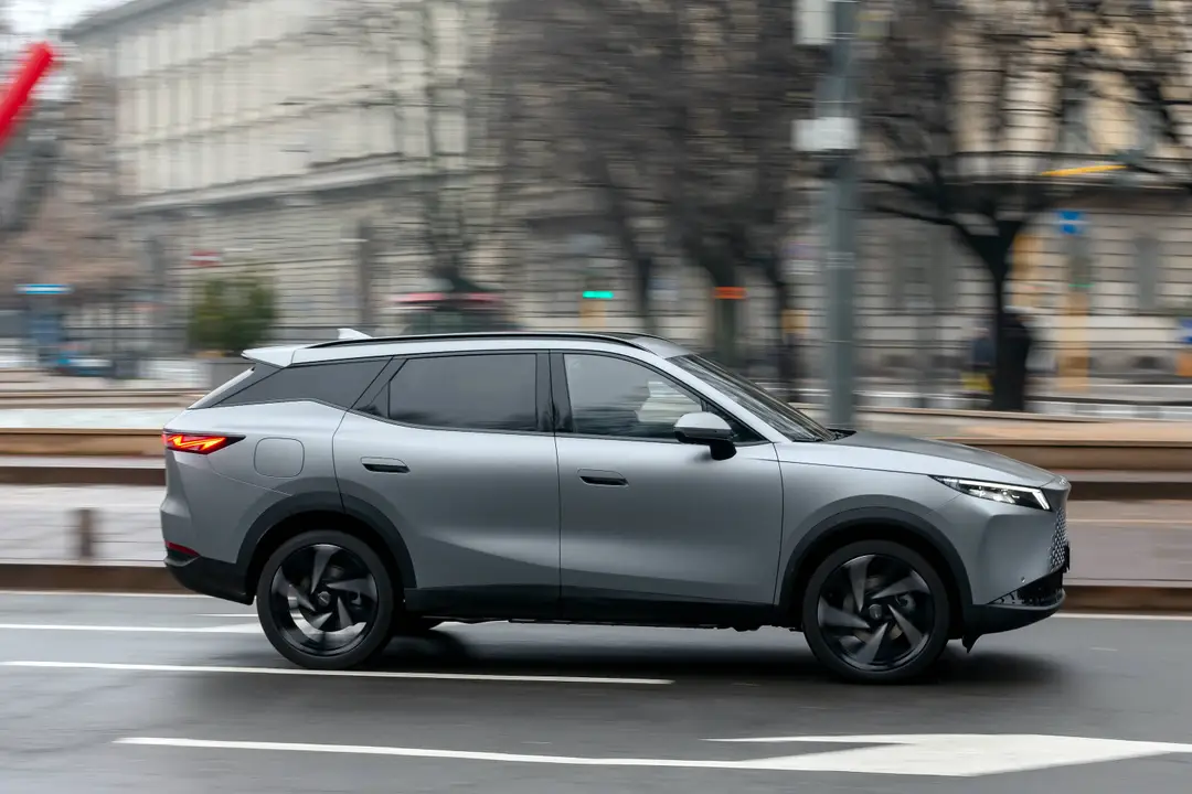 Scatto dinamico laterale (panning) di Omoda 7 SHS-P in movimento. L'effetto di velocità sullo sfondo urbano evidenzia la fluidità di marcia e il design aerodinamico del nuovo SUV ibrido plug-in.