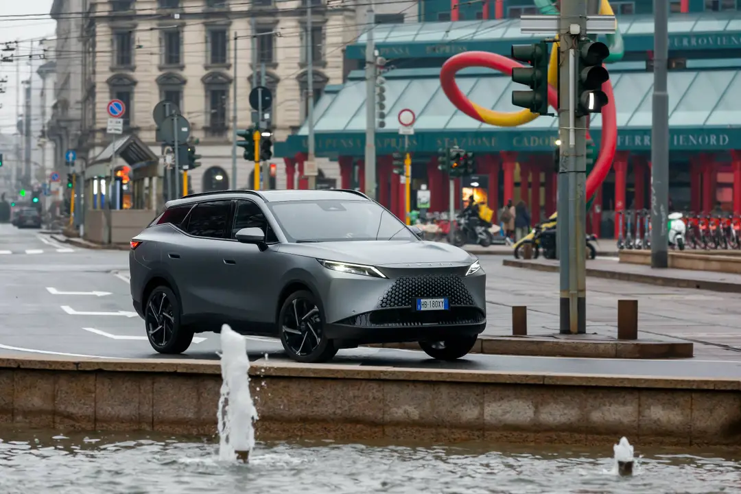 Omoda 7 SHS-P in movimento nel centro di Milano, zona Cadorna. Il SUV ibrido plug-in in colorazione Matte Soft Grey transita su strada urbana con la scultura "Ago, Filo e Nodo" visibile sullo sfondo.