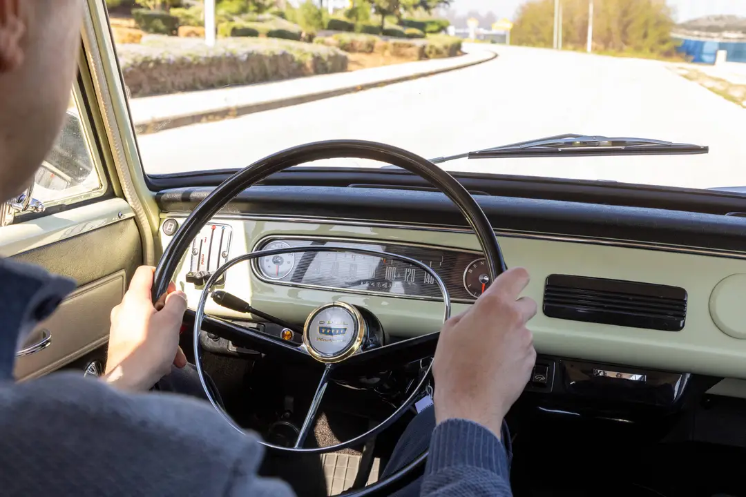 Federico Ferrero view inside a vintage Opel Kadett. Hands grip a thin steering wheel with a chrome ring, facing a cream-colored analog dashboard and road
