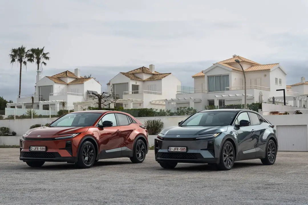 Foto di due esemplari di Toyota C-HR+, una rossa metallizzata e l'altra grigia metallizzata, fotografate frontalmente con una città marittima di sfondo