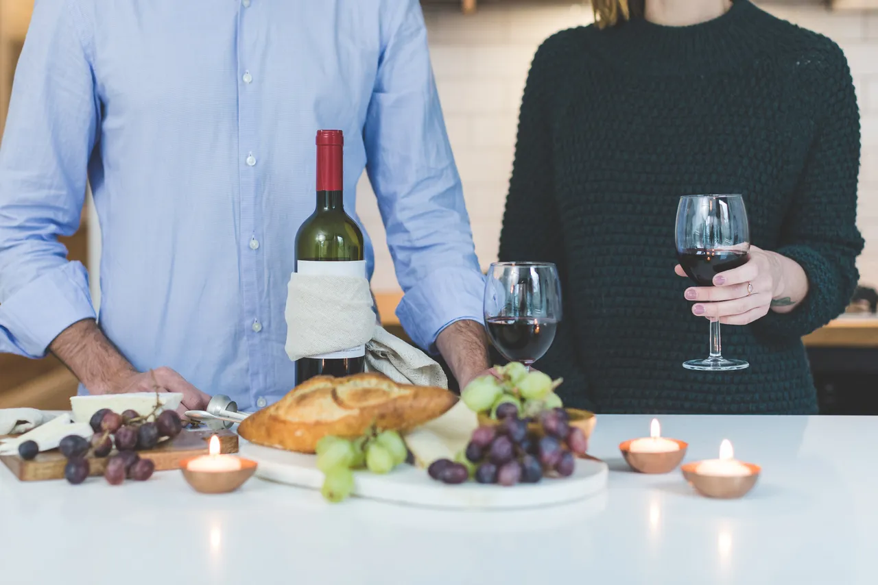 canva man standing beside woman holding wine glass in front of grapes and bread on table