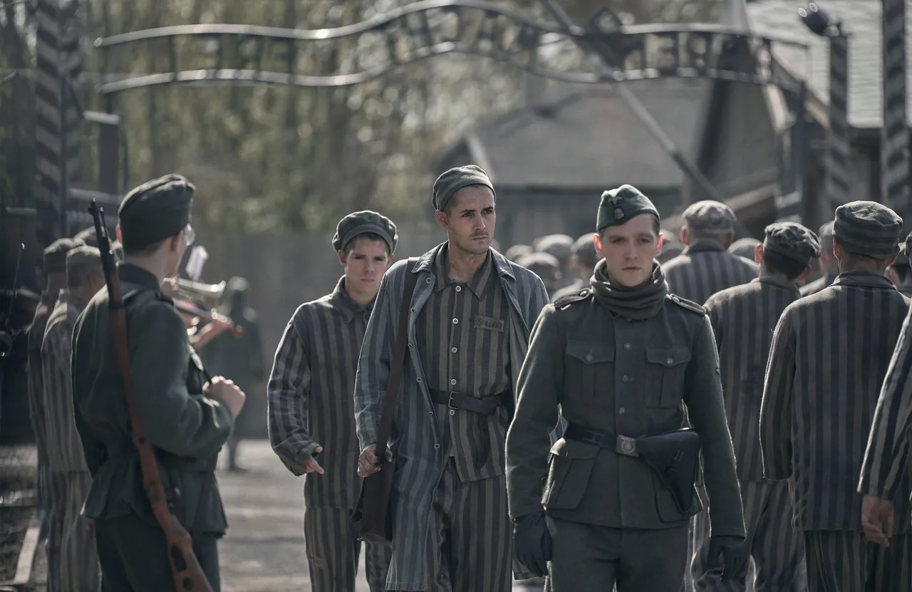 Caption: Jonah Hauer-King as Lali Sokolov seen here walking through Auschwitz with German-born actor Jonas Nay as Nazi officer Stefan Baretzki, front right (Credit: © Martin Mlaka / SkyShowtime)