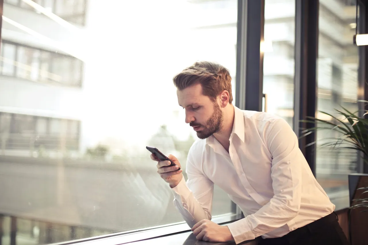 photo of man in white dress shirt holding phone near window 859265f1595921456