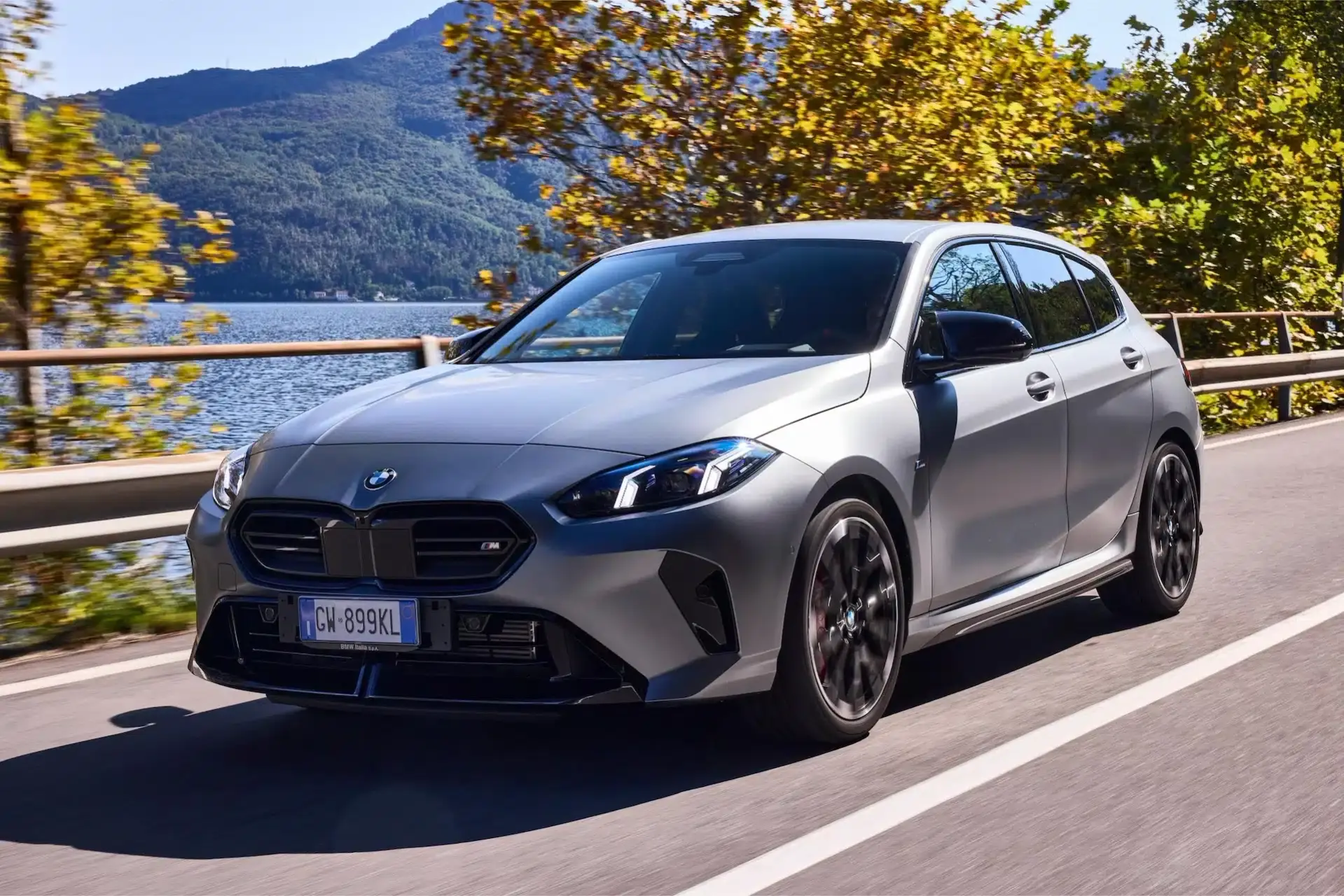 bmw serie 1 2024 di colore grigio che avanza su una strada con un lago sullo sfondo