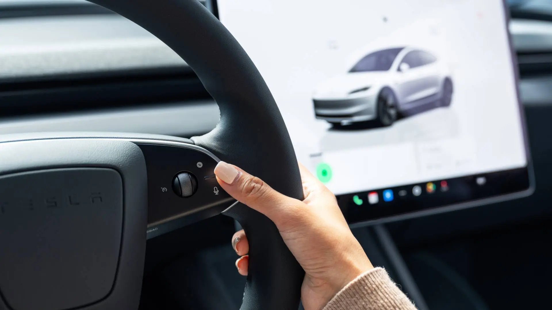 Volante della Tesla Model 3 in primo piano con un dito che tocca il tasto con il microfono e sul fondo lo schermo della model  che mostra il veicolo da fuori