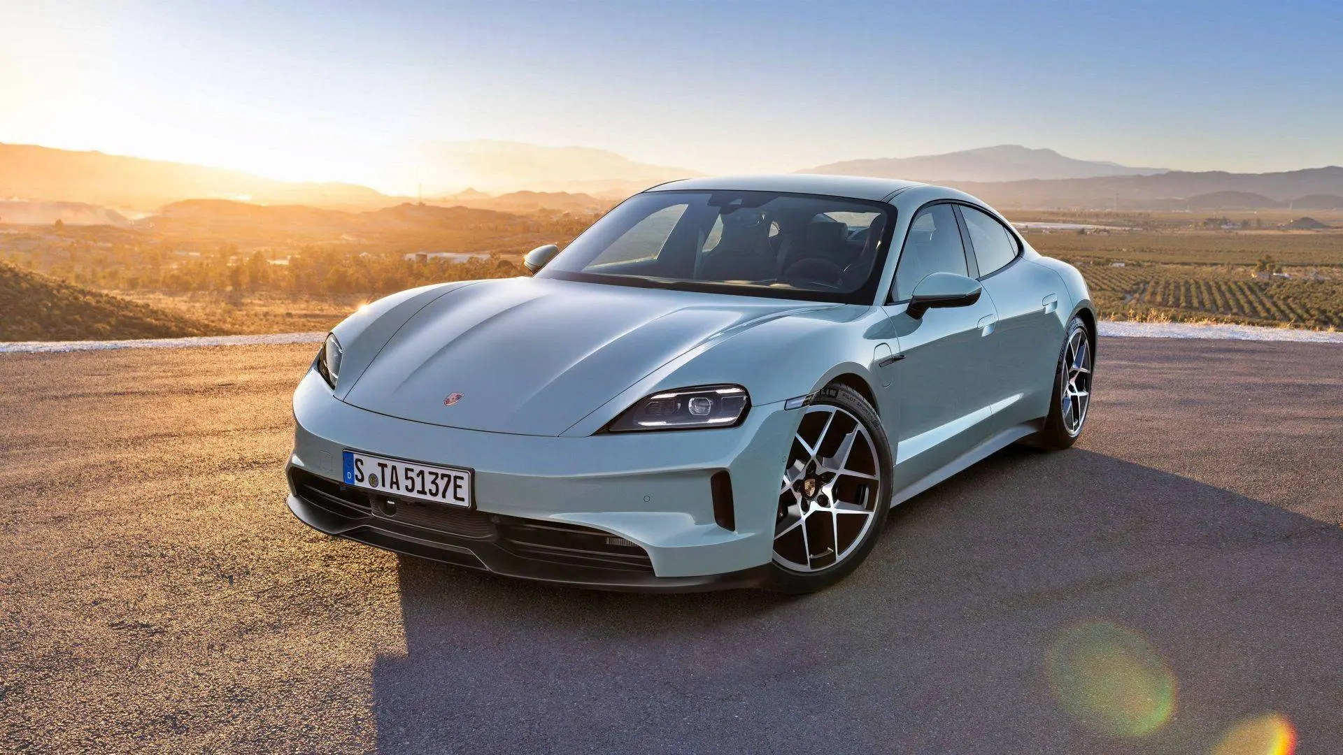 Nell'immagine si vede una Porsche Taycan grigia su uno sfondo collinare al tramonto