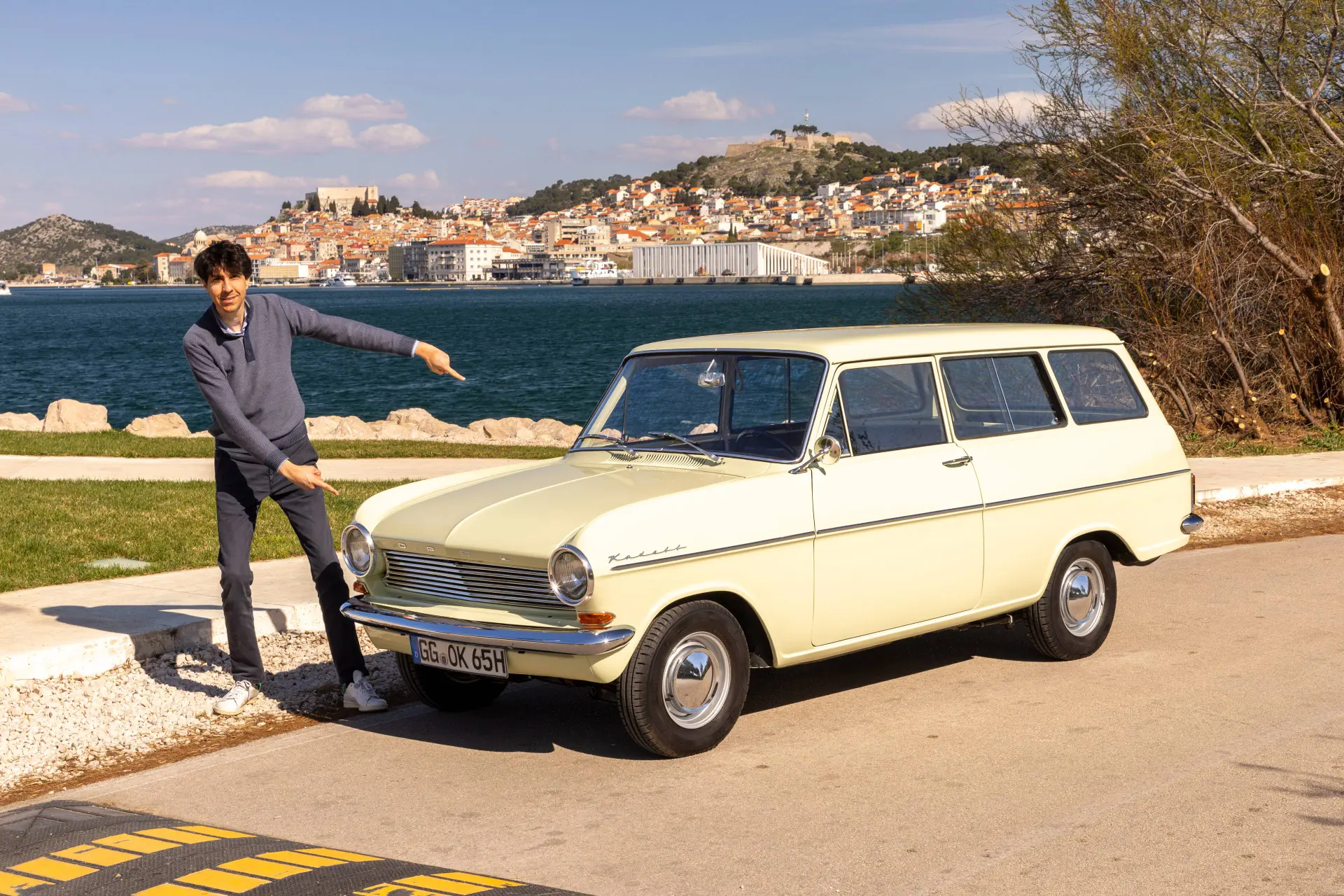 Federico Ferrero indica una rarissima Opel Kadett A Caravan storica del 1965, di colore crema chiaro, parcheggiata sul lungomare asfaltato di Mandalina, in Croazia. Lo sfondo mostra un porto e una città collinare con una fortezza sotto un cielo sereno