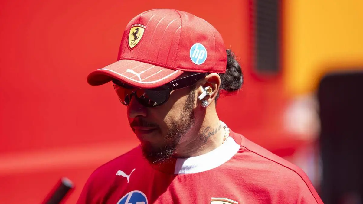 Lewis Hamilton voor een rode achtergrond met een Ferraripetje op.
