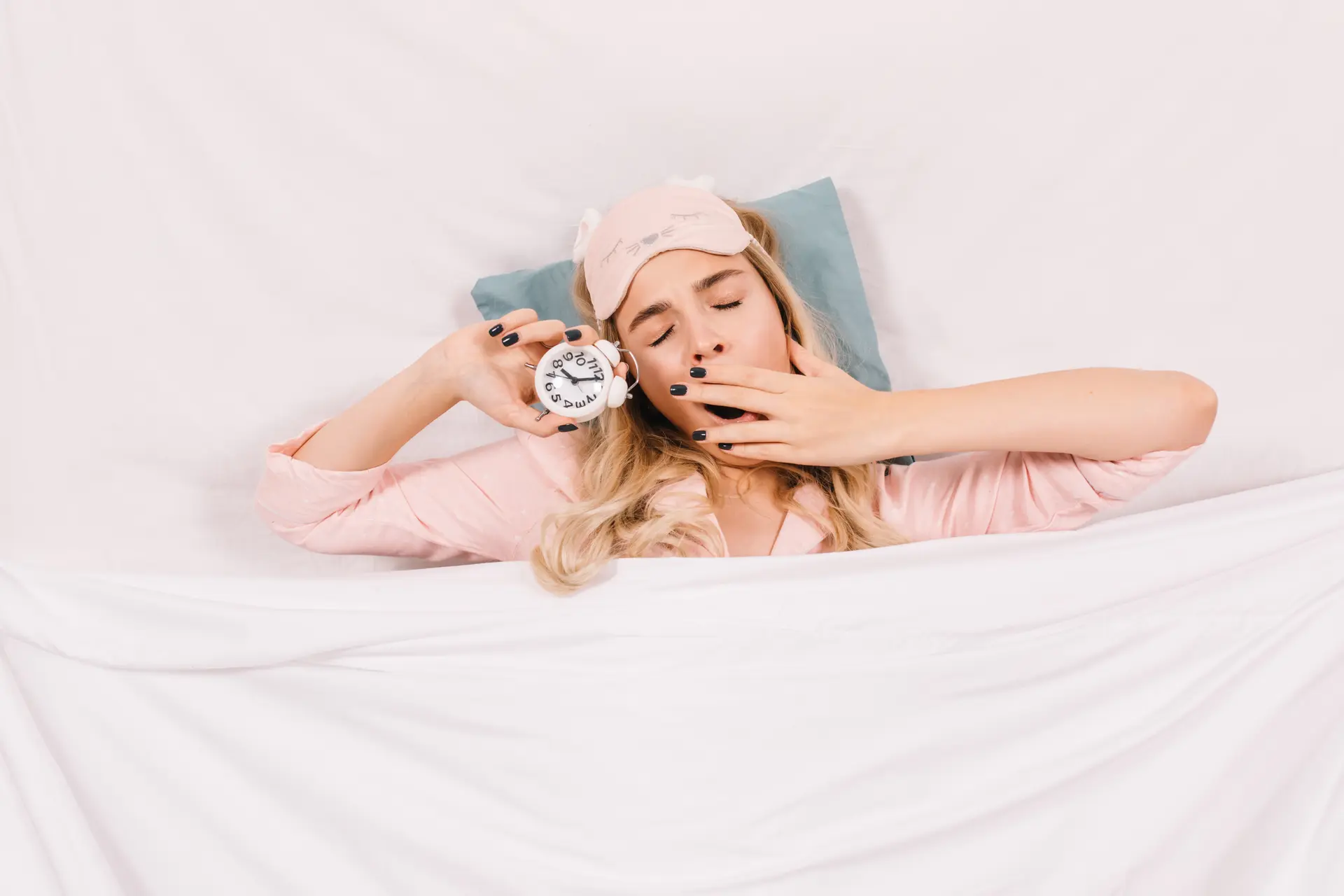 Top view of sleepy girl yawning in bed. Indoor shot of caucasian young woman posing with clock