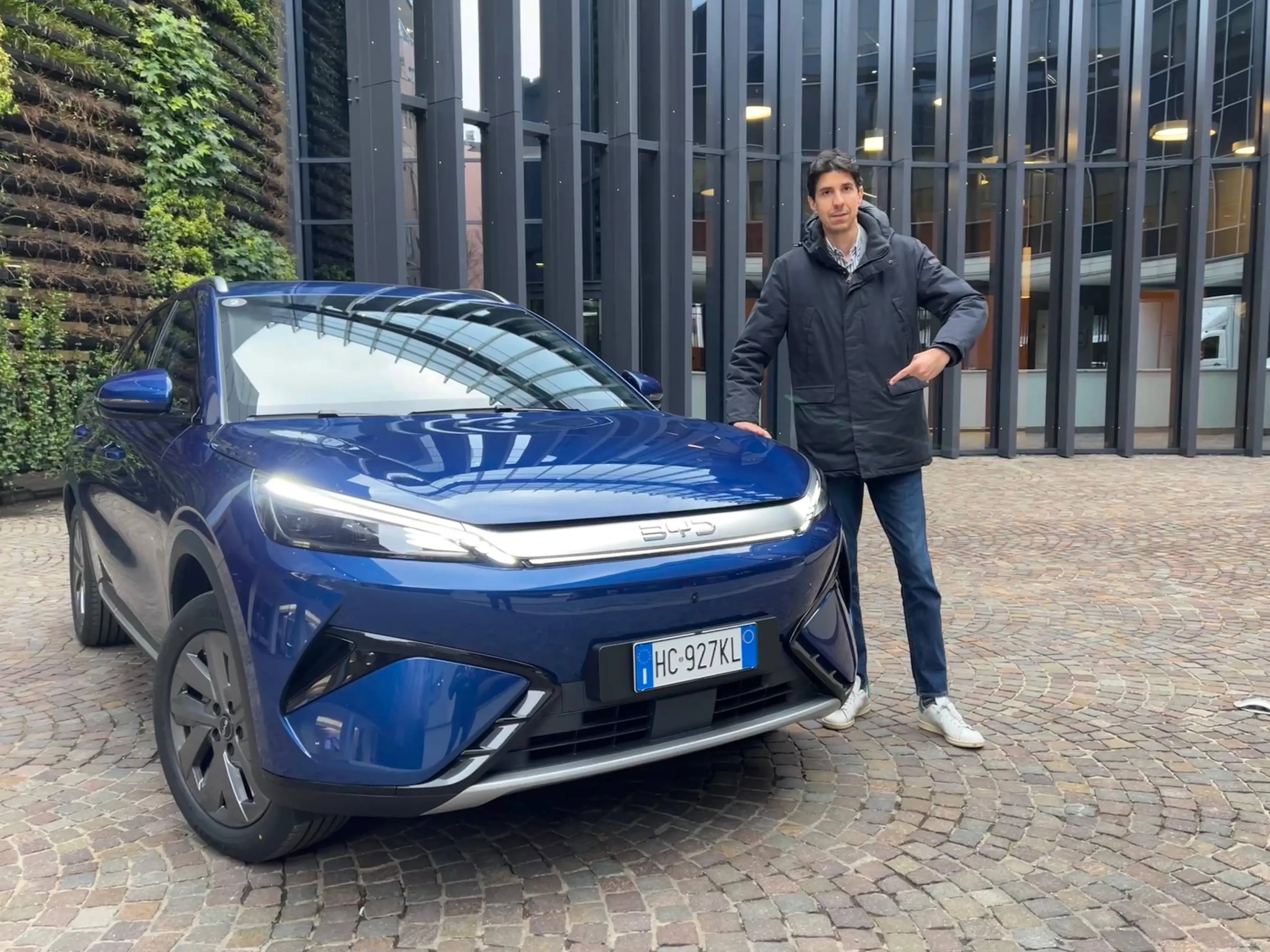 Federico Ferrero vicino alla BYD Atto 3 EVO di colore blu scuro, mentre indica il frontale dell'auto. La vettura è parcheggiata su una pavimentazione in pietra davanti a un edificio moderno con una parete di verde verticale