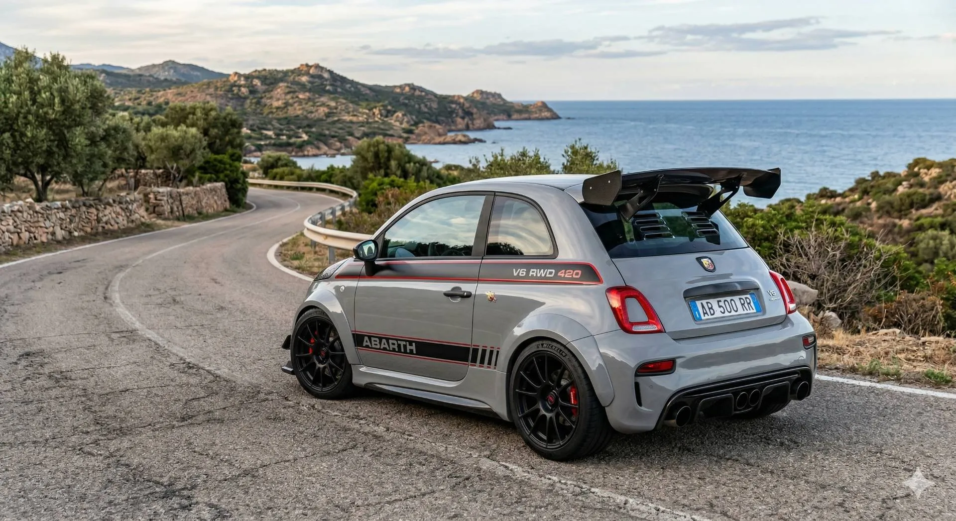 Una vista posteriore a tre quarti di una Abarth 500 ampiamente personalizzata e performante, di colore grigio scuro con finitura satinata, ferma su una strada asfaltata sinuosa che costeggia il mare e colline rocciose mediterranee.