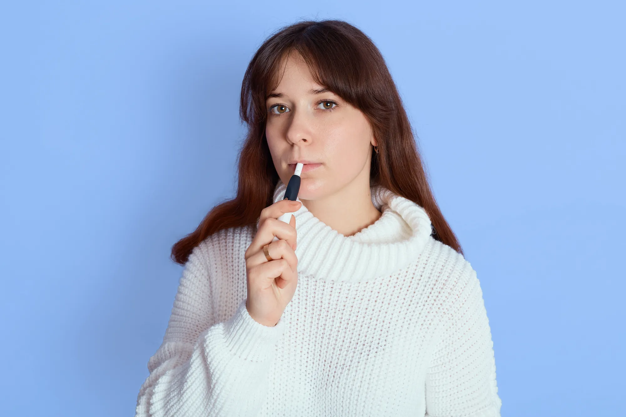 young pretty girl with electronic cigarette posing over blue looks at camera wearing white sweater dark haired female smoking e cigarette indoor