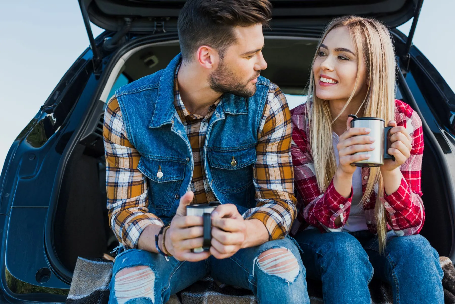 Couple of Young Tourists With Coffee Cups Sitting on Car Trunk