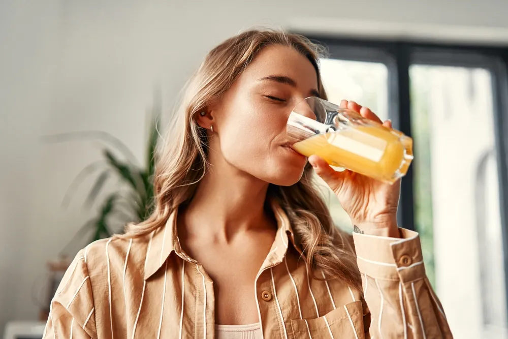 A,Young,Happy,Woman,Standing,In,The,Kitchen,Drinks,Orange