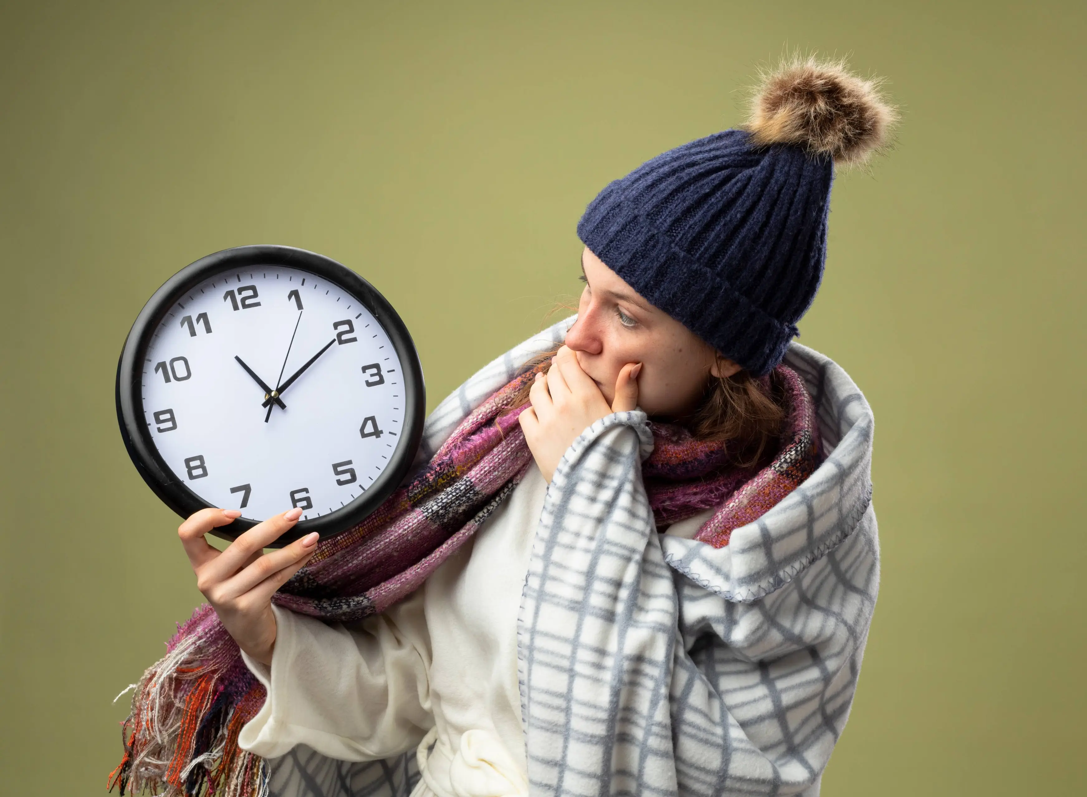 scared-young-ill-girl-wearing-white-robe-winter-hat-with-scarf-holding-looking-wall-clock-wrapped-plaid-isolated-olive-green