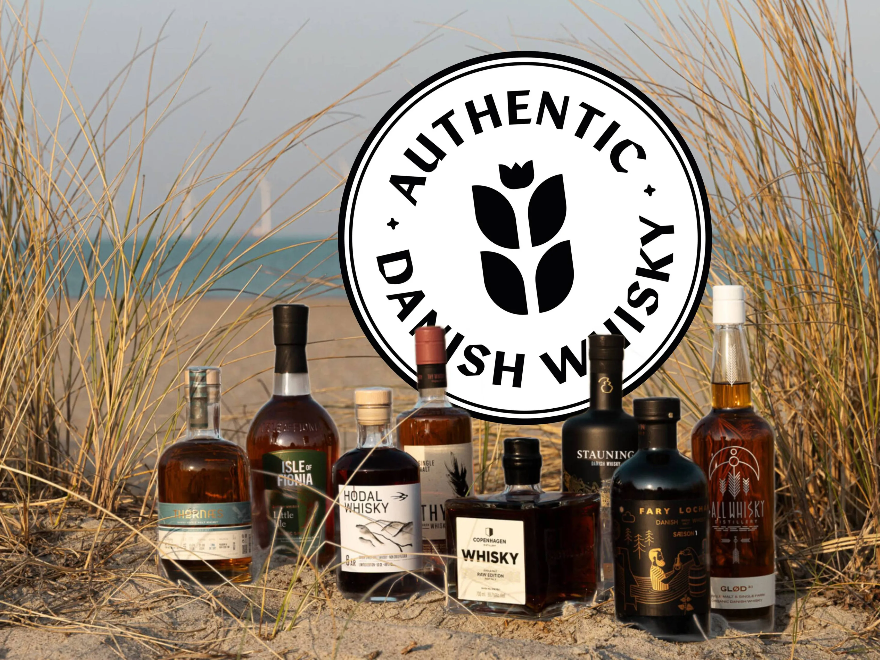 Deense whiskyflessen op een strand met duingras en het Authentic Danish Whisky logo