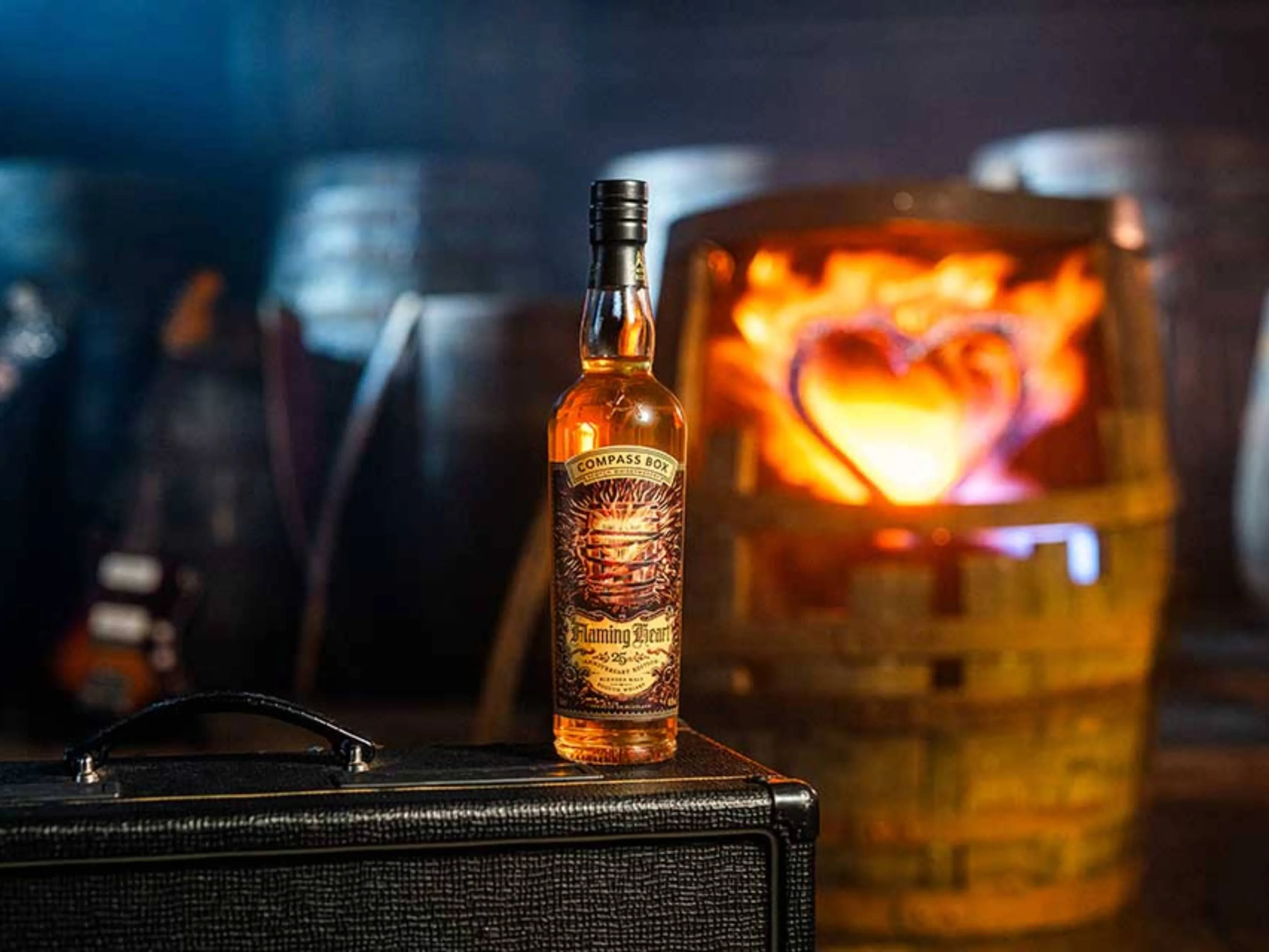 Een fles Compass Box Flaming heart 2025 25th Anniversary Edition whisky met op de achtergrond een brandend vat met een hart erin