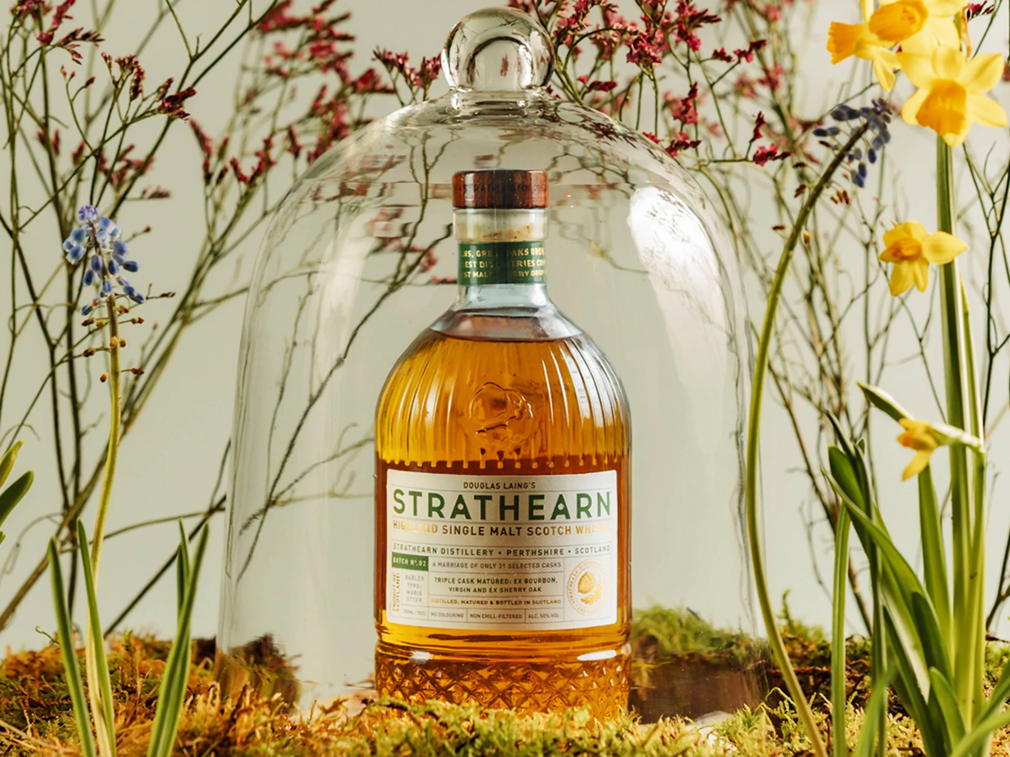 Een fles Strathearn Batch 2 whisky onder een stolp bij lentebloemen
