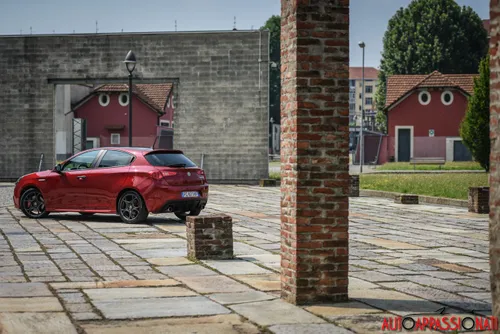 Foto di una Alfa Romeo Giulietta Veloce fotografata di tre quarti posteriore, di colore Rosso Competizione e parcheggiata in una corte con una colonna di mattoni sul lato destro.