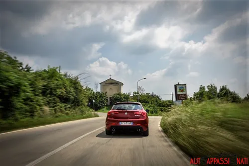 Foto di una Alfa Romeo Giulietta Veloce dinamica, con ripresa del posteriore pieno su una strada di campagna con alberi e una chiesetta davanti alla macchina.