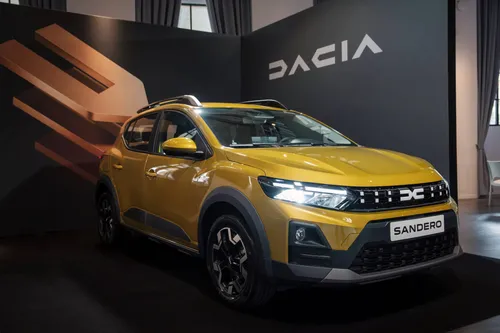 Una Dacia Sandero Stepway 2026di colore giallo dorato metallizzato esposta in uno showroom. L'auto è ripresa di tre quarti anteriore e mostra la nuova calandra con il logo Dacia bianco, fari a LED accesi e barre sul tetto.
