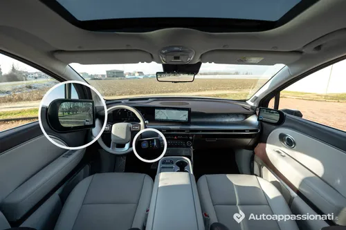 Panoramica degli interni della Hyundai IONIQ 9: i cerchi bianchi evidenziano il display dello specchietto laterale digitale e il selettore del cambio shift-by-wire sul piantone dello sterzo.