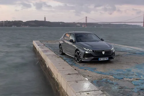 il lato anteriore della Peugeot 308 2026 di tre quarti, parcheggiata sulla riva del fiume Tago a Lisbona, con dietro il Ponte 25 Aprile