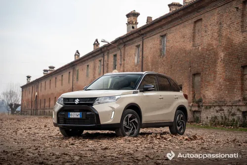 Foto frontale di tre quarti della Suzuki Vitara Hybrid AllGrip 2026 parcheggiata su fogliame e con dietro una cascina