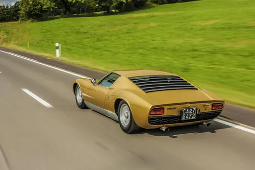 Esemplare di Lamborghini Miura S di colore ocra ripresa dal posteriore che percorre strada con prato sullo sfondo