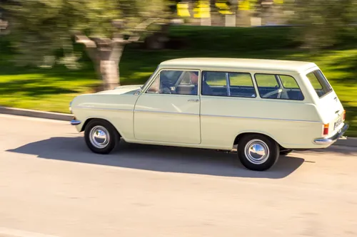 Dynamic side view of a vintage cream-colored Opel Kadett Caravan A driving on a road with a motion-blurred background of grass and trees