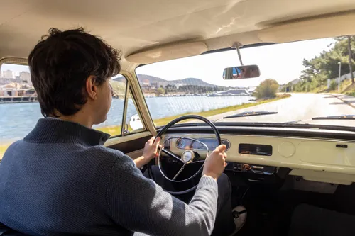 Federico Ferrero driving Opel Kadett Caravan viewed from behind. Through the windshield: a winding coastal road, water, and a town.