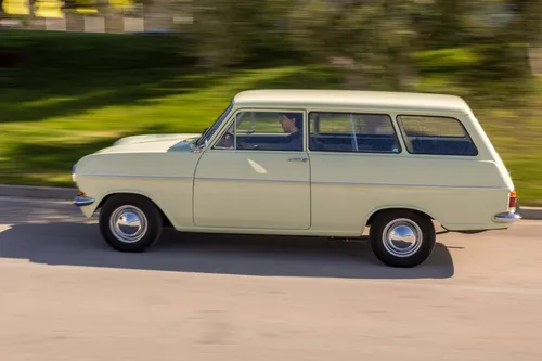 Dynamic side view of a vintage cream-colored Opel Kadett Caravan A driving on a road with a motion-blurred background of grass and trees.