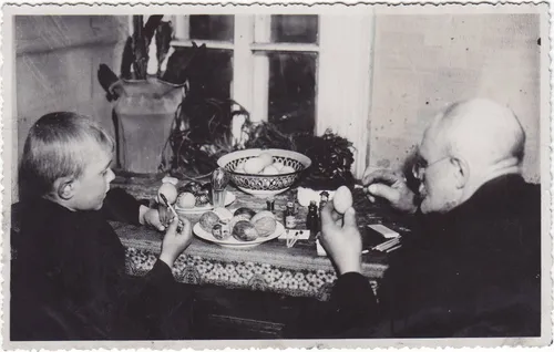 Fotonuotrauka. Grupinė. Senelis (dešinėje) su anūku (kairėje) dažo velykinius margučius prie uždaroje patalpoje esančio stalo. XX a. 3–4 deš. (Trakų muziejus, CC BY)