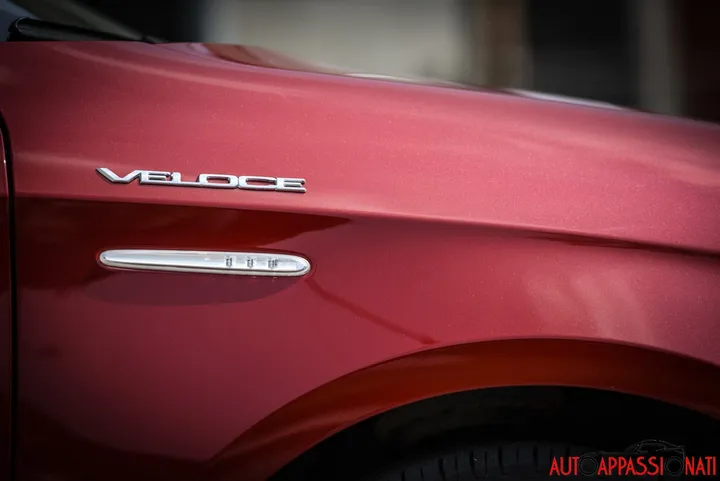Foto del dettaglio del logo Veloce dell'Alfa Romeo Giulietta più sportiva, con il logo cromato sopra la freccia laterale sulla carrozzeria verniciata in colore Rosso Competizione.