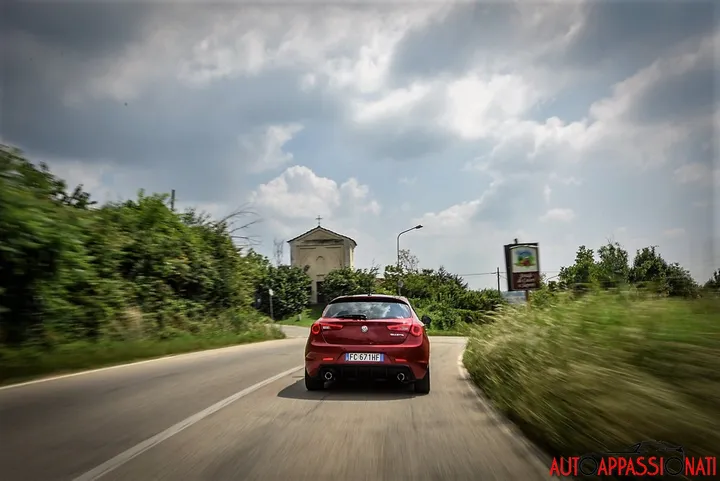Foto di una Alfa Romeo Giulietta Veloce dinamica, con ripresa del posteriore pieno su una strada di campagna con alberi e una chiesetta davanti alla macchina.