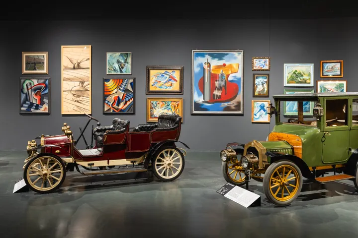 Una sala espositiva con pareti scure su cui sono appesi dipinti futuristi, con due automobili d'epoca parcheggiate in primo piano esposte al Museo dell'auto di Torino