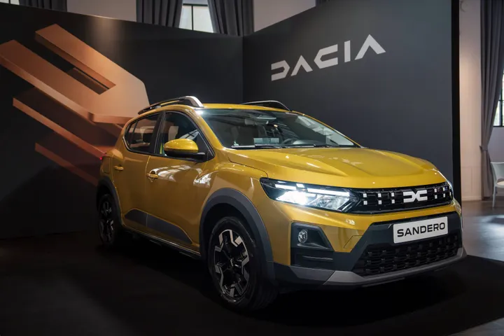 Una Dacia Sandero Stepway 2026di colore giallo dorato metallizzato esposta in uno showroom. L'auto è ripresa di tre quarti anteriore e mostra la nuova calandra con il logo Dacia bianco, fari a LED accesi e barre sul tetto.
