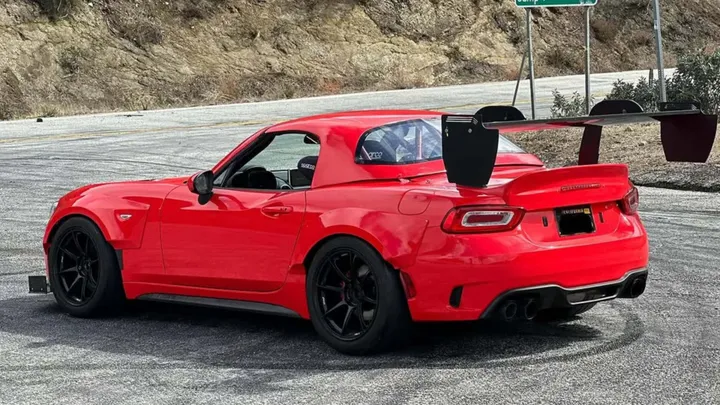 Nella foto si vede una 124 spider rossa di profilo con un alettone nero di grandi dimensioni. La foto è stata scattata in una piazzola di sosta di una strada collinare americana