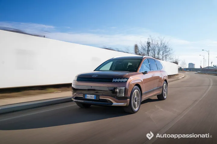 Hyundai Ioniq 9 bronzo opaco in movimento su strada soleggiata. Ripresa di tre quarti che evidenzia i fari LED a 'H' e la griglia. Lo sfondo con muro bianco sfocato enfatizza la velocità del veicolo.