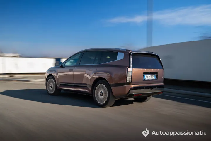Vista posteriore di tre quarti di una Hyundai Ioniq 9 color bronzo opaco in movimento su strada. L'immagine evidenzia il design squadrato del retro, l'ampio portellone verticale e i fanali posteriori posizionati in basso.