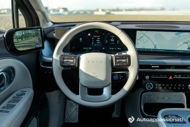 Vista completa del posto di guida della Hyundai Ioniq 9, che include il volante multifunzione a due razze, il doppio display digitale curvo per strumentazione e infotainment, e la console centrale con i comandi del climatizzatore.