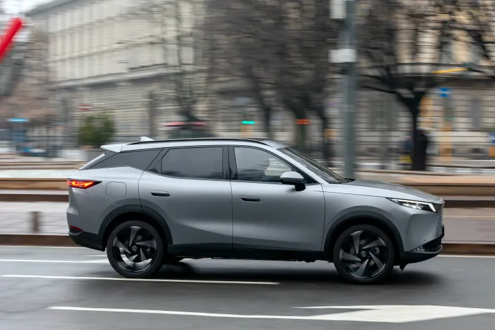 Scatto dinamico laterale (panning) di Omoda 7 SHS-P in movimento. L'effetto di velocità sullo sfondo urbano evidenzia la fluidità di marcia e il design aerodinamico del nuovo SUV ibrido plug-in.