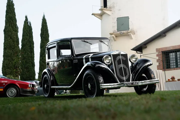 Foto a colori di una Lancia Augusta restaurata, con il numero 6 verniciato sulla griglia e targa originale 7033-PA, esposta su un prato verde con un edificio rurale e cipressi sullo sfondo.