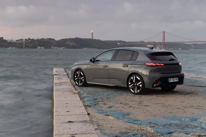 il lato posteriore della Peugeot 308 2026 di tre quarti, parcheggiata sulla riva del fiume Tago a Lisbona, con dietro il Ponte 25 Aprile