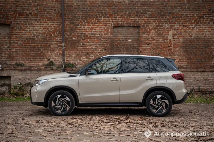 Foto laterale della Suzuki Vitara Hybrid AllGrip 2026, parcheggiata su fogliame e con un muro di mattoni sullo sfondo