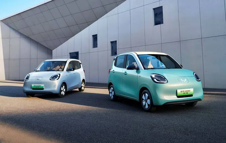 Due Wuling Mini EV parcheggiate davanti a un edificio moderno geometrico. A sinistra l'auto è azzurra, a destra è verde menta con tetto bianco. Entrambe mostrano il design squadrato e compatto tipico di questa popolare city car elettrica cinese.
