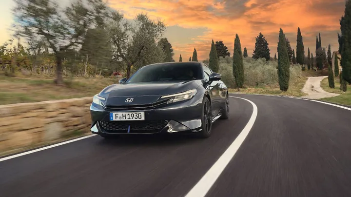 Una foto dinamica dell'anteriore della Honda Prelude Hybrid 2026, verniciata in colore grigio e con un tramonto e delle strade collinari toscane