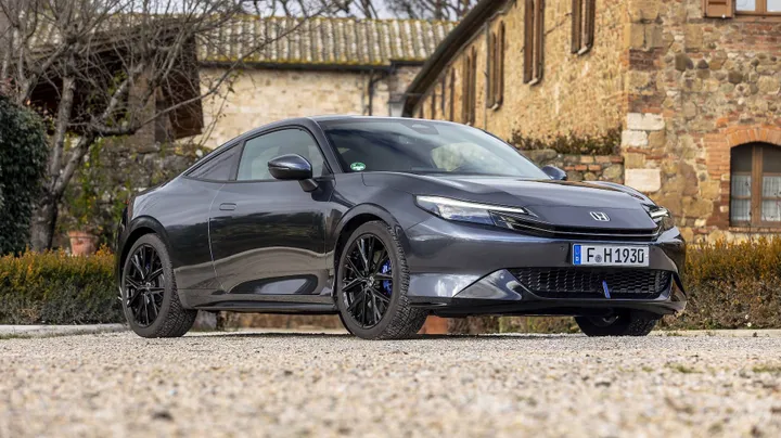 Una foto della Honda Prelude Hybrid 2026 dal basso, di tre quarti anteriore, di colore grigio e con, dietro, un casolare d'epoca
