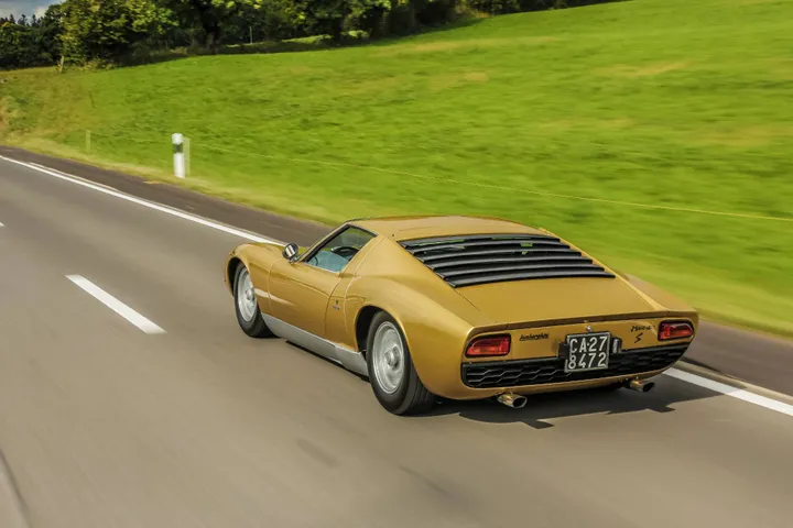 Esemplare di Lamborghini Miura S di colore ocra ripresa dal posteriore che percorre strada con prato sullo sfondo