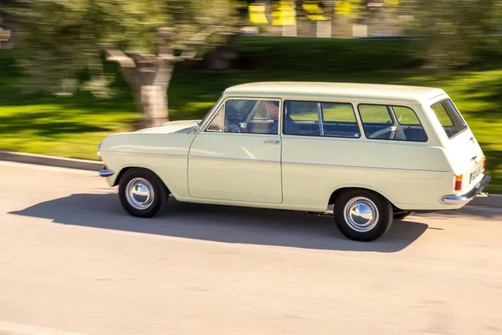 Dynamic side view of a vintage cream-colored Opel Kadett Caravan A driving on a road with a motion-blurred background of grass and trees